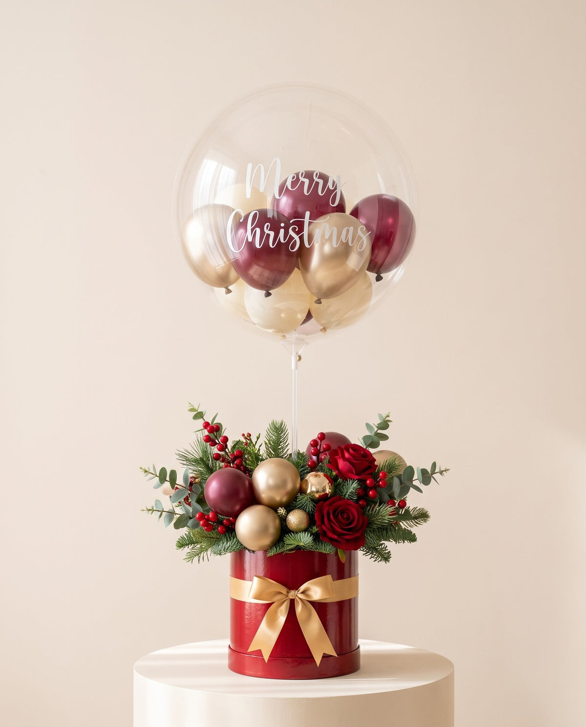 Clear bubble balloon lettered Merry Christmas, filled with burgundy, gold and ivory mini balloons, rising from a red hamper bucket with festive pine greenery, red berries and gold accents, on a cream plinth (design concept)