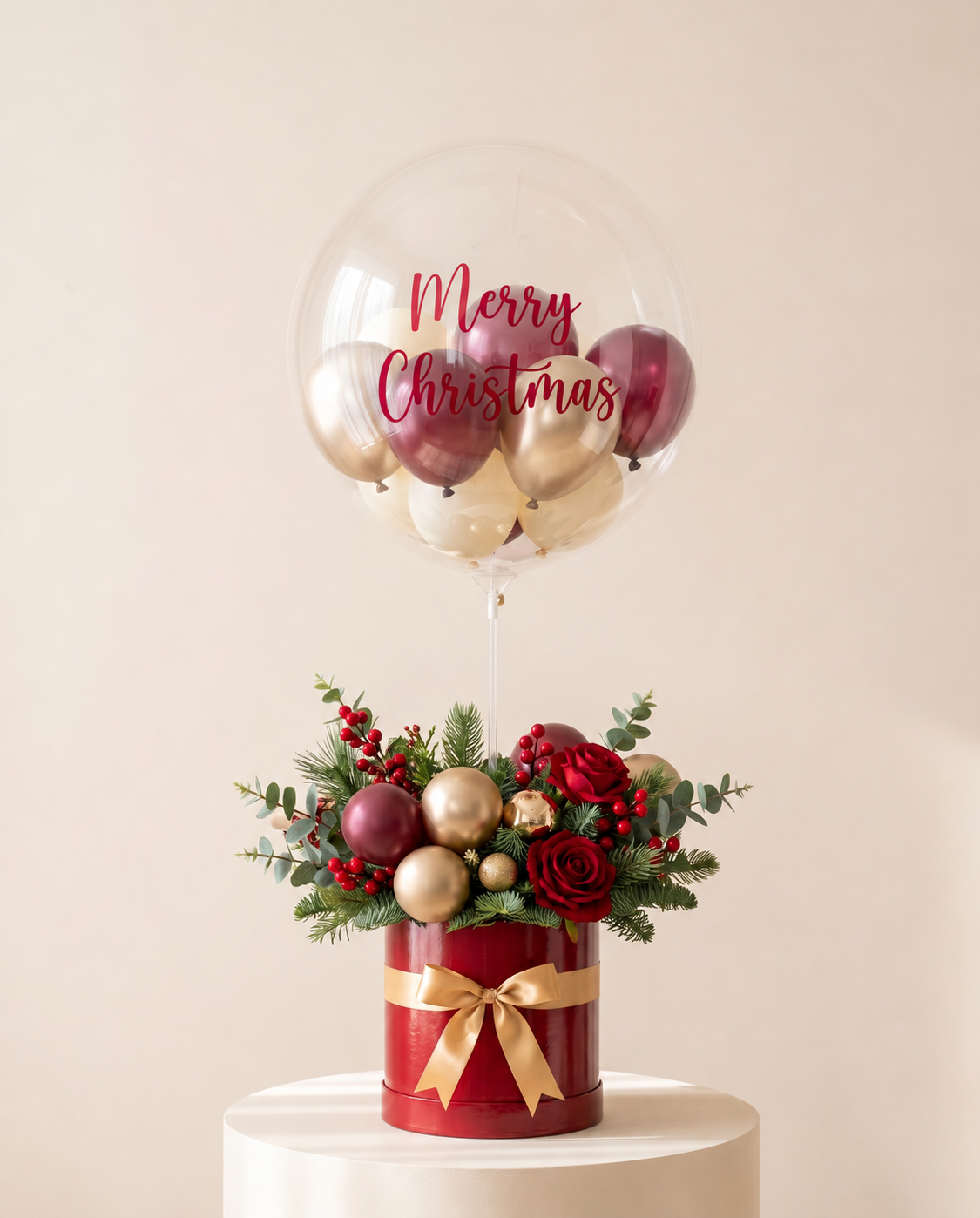 Clear bubble balloon lettered Merry Christmas in red script, filled with burgundy, gold and ivory mini balloons, above a red hat box of pine, berries and red roses with a gold bow (design concept). Red lettering; main view.