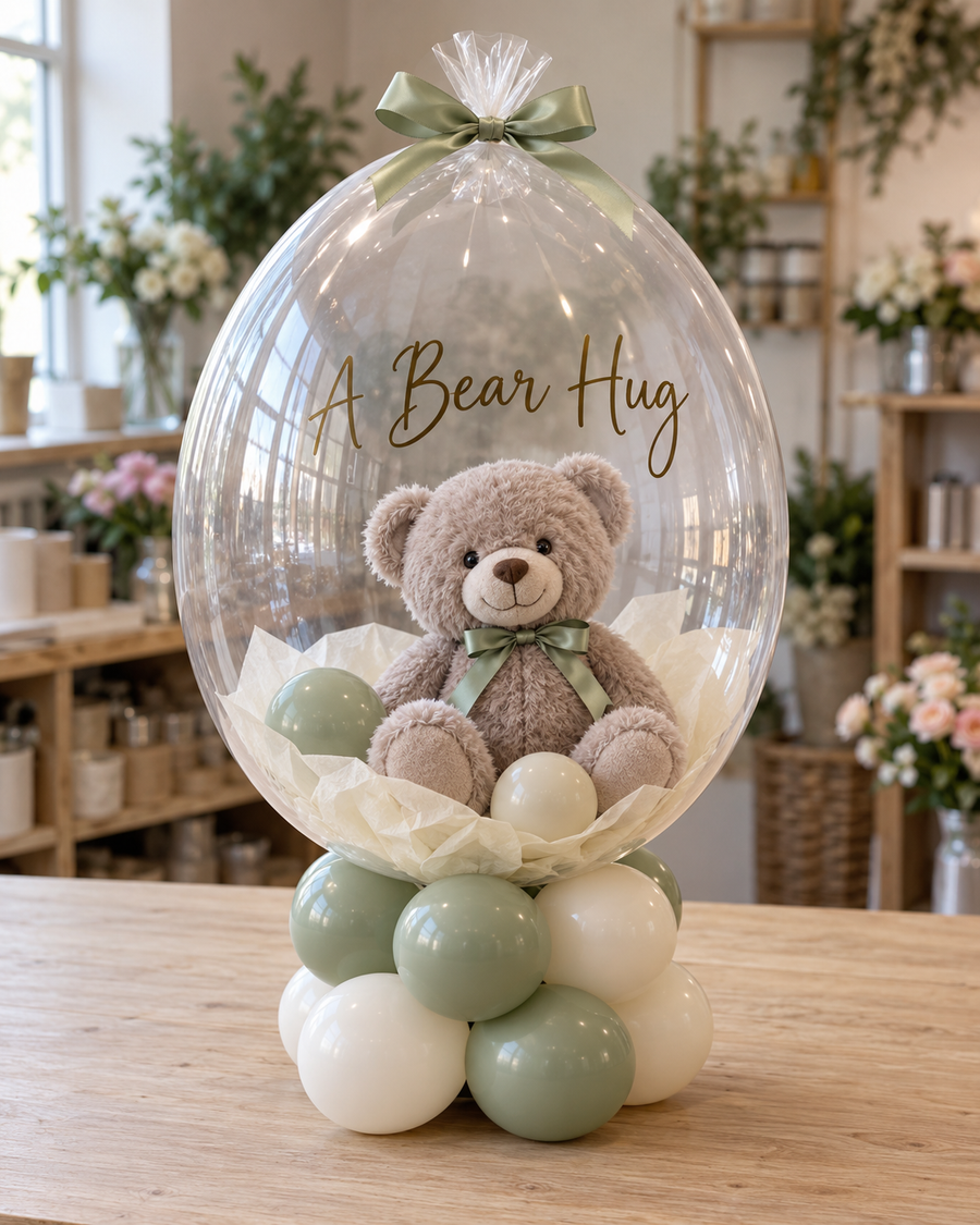 Gift studio original concept 20261009: A Bear in a Bubble. A soft teddy enclosed inside a clear gift balloon, sitting on a sage and ivory balloon base. AI concept; construction subject to prototype.