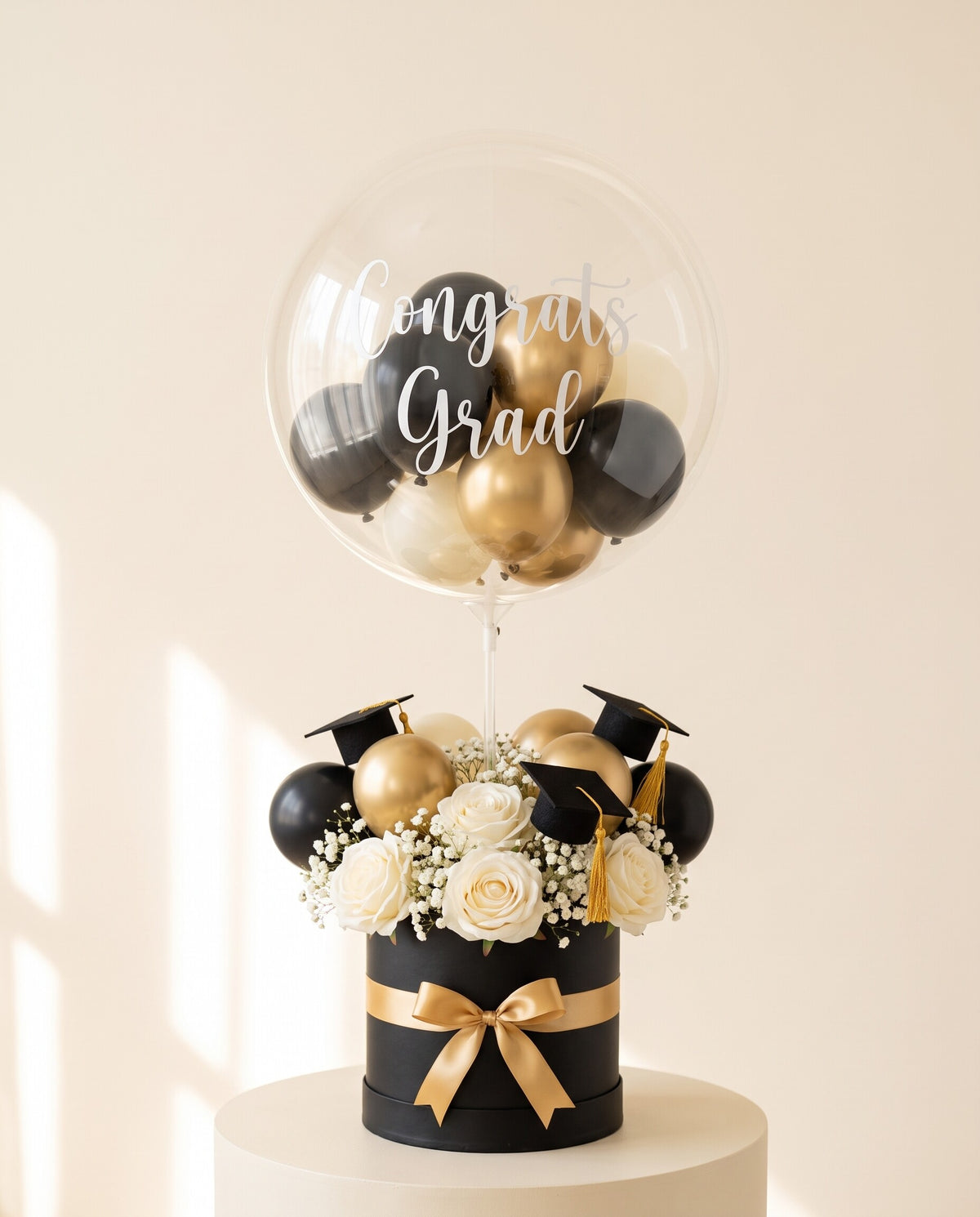 Clear bubble balloon lettered Congrats Grad, filled with black and gold mini balloons, above a black round hat box with ivory roses, gold accents and three small black mortarboard toppers, tied with gold satin ribbon, on a cream plinth (design concept)