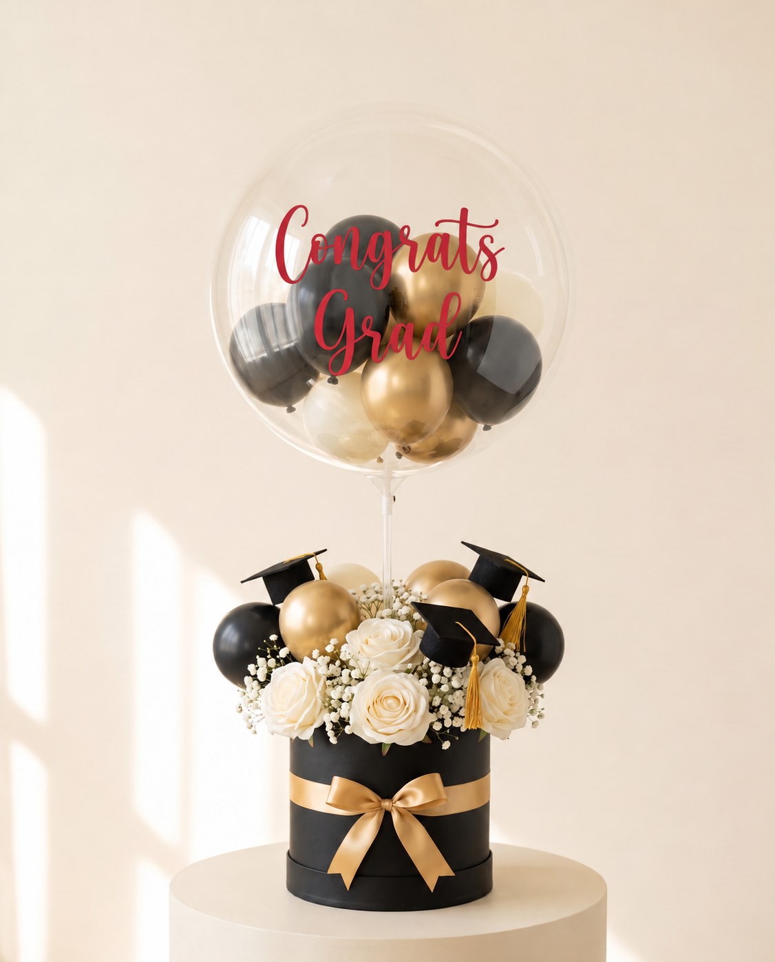 Clear bubble balloon lettered Congrats Grad in red script, filled with black and gold mini balloons, on a black hat box of ivory roses with graduation cap toppers and a gold bow (design concept)